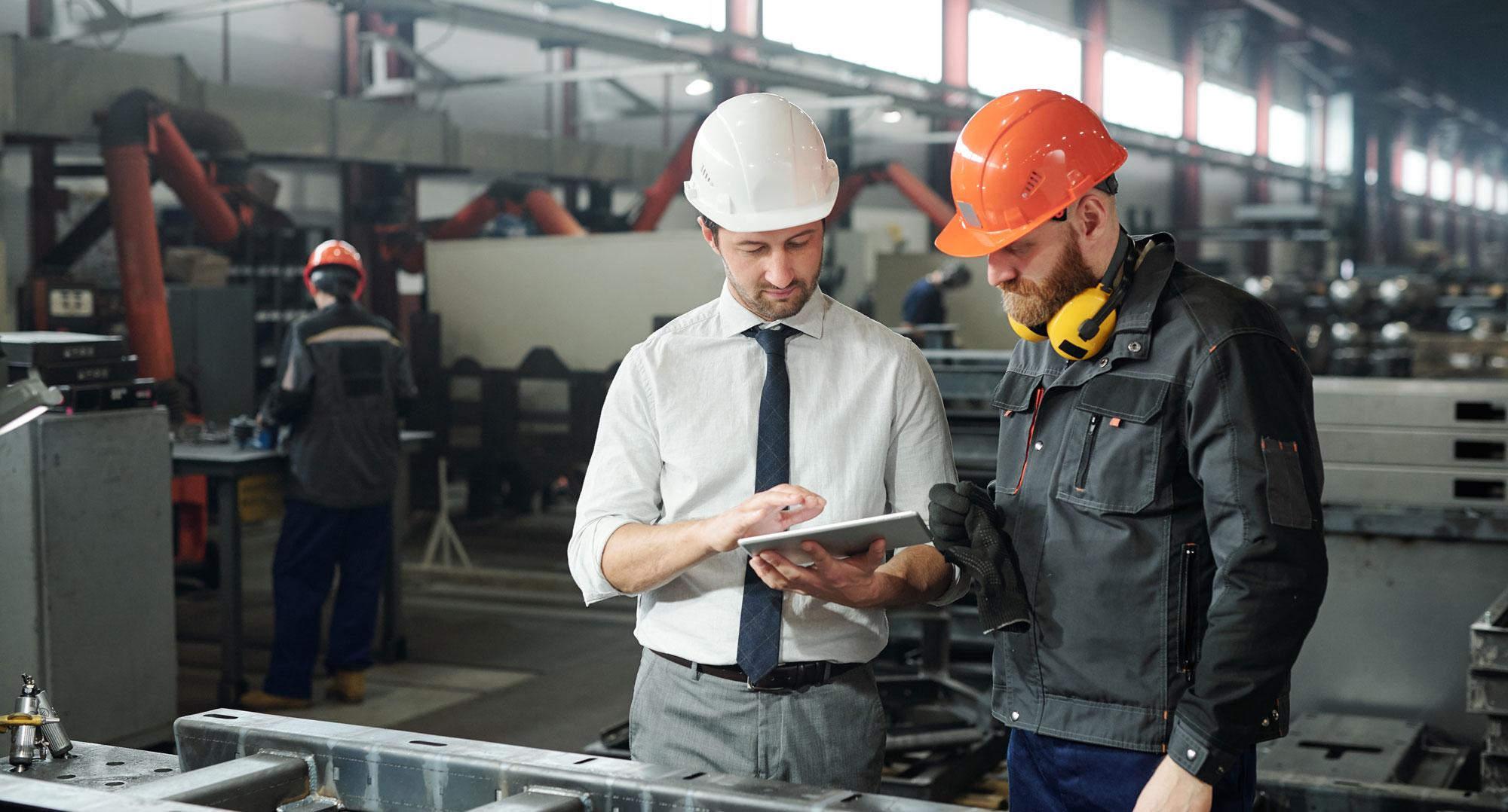 Two men in a factory wearing hard hats and viewing a tablet
