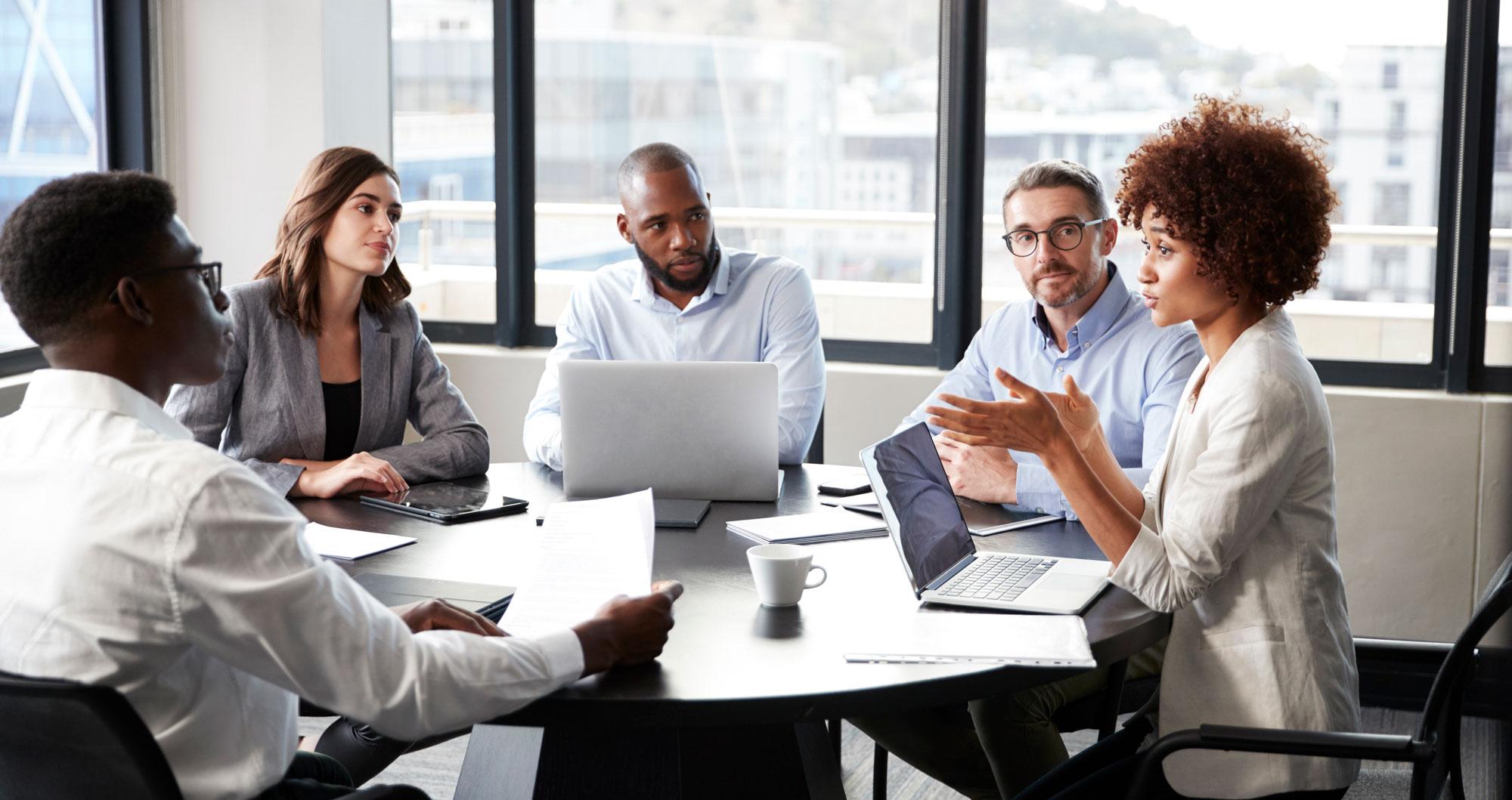 Five people sitting in a meeting with laptops and tablets